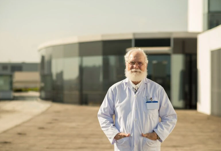 An old man with a white beard standing in front of a building equipped with solar panels.