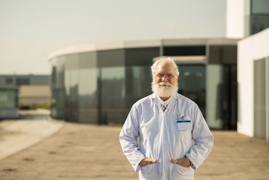 An old man with a white beard standing in front of a building equipped with solar panels.