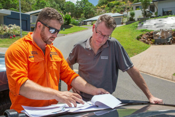 Two men in orange shirts looking at paperwork on the hood of a car discussing solar power options.
