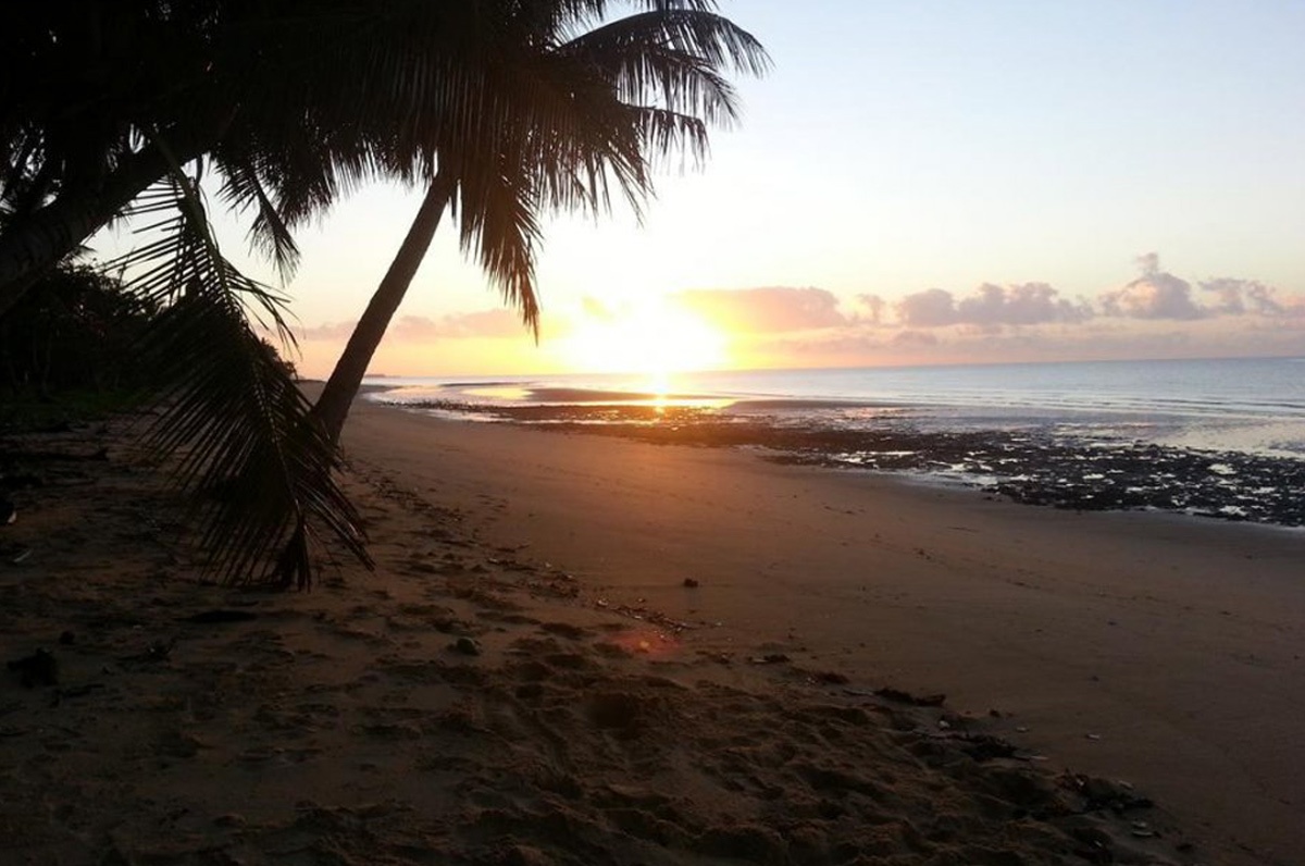 The sun is setting over a sandy beach with palm trees, casting a beautiful glow on the Cairns.