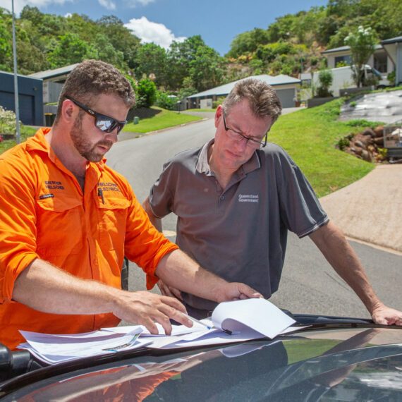 Two men in orange shirts, affiliated with Hielscher Electrical, are considering paperwork related to solar power on the hood of a car.