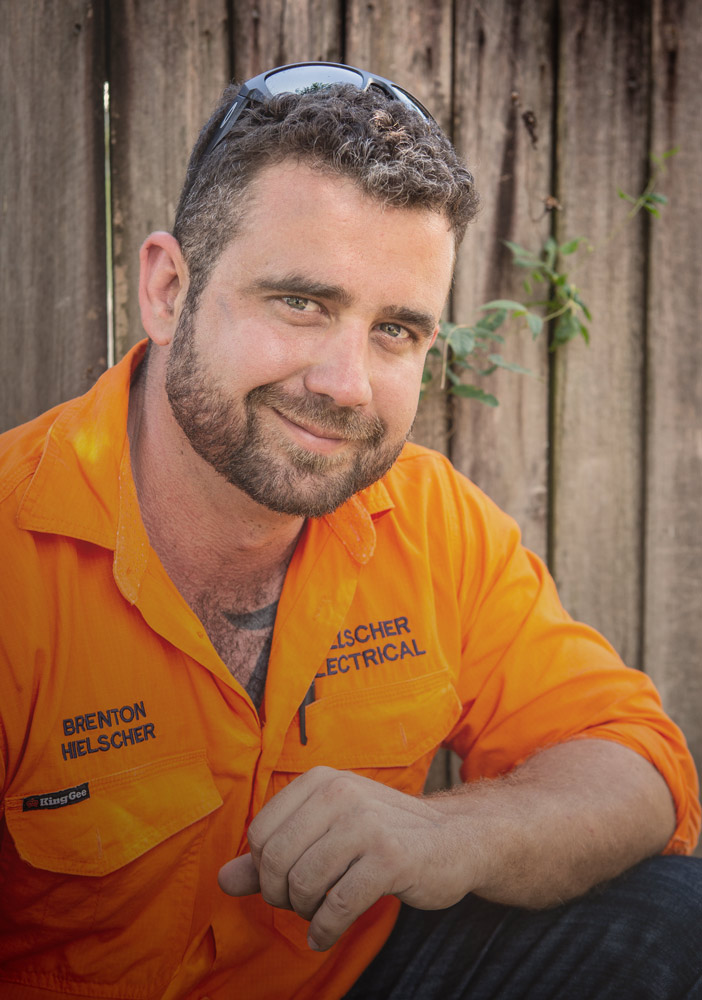 Brenton Hielscher in an orange shirt leaning against a fence while Hielscher Electrical installs solar panels nearby.