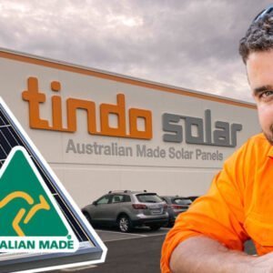 A man in an orange shirt is standing next to a solar panel, harnessing solar power.