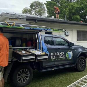 Two men, hired by Hielscher Electrical, are installing solar panels on the roof of a house, harnessing the power of the sun through Solar Power.