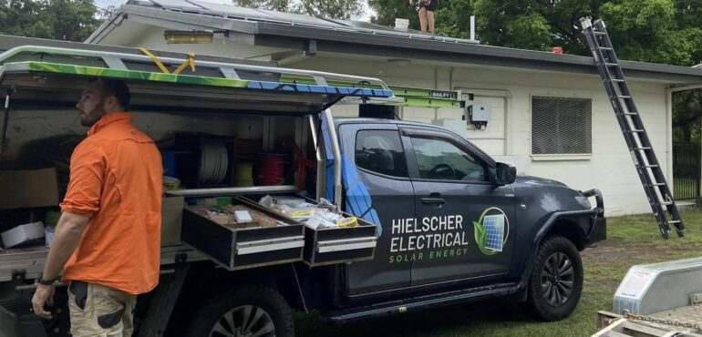 Two men, hired by Hielscher Electrical, are installing solar panels on the roof of a house, harnessing the power of the sun through Solar Power.