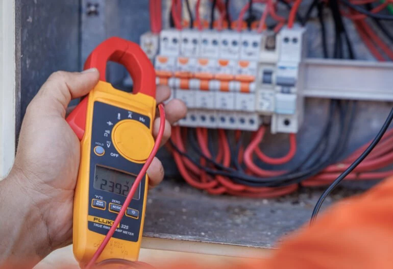A person from Hielscher Electrical, in Cairns, holding a multimeter in front of an electrical panel.
