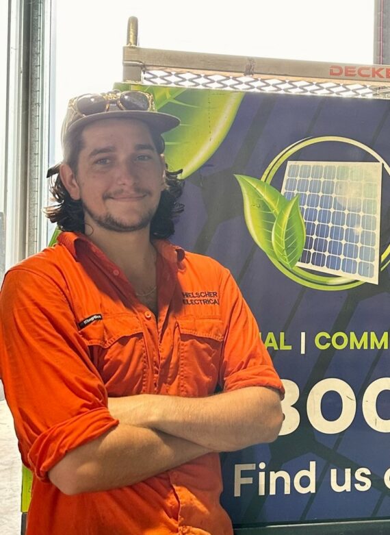 A young solar electrician named Harry in an orange work shirt and a cap stands smiling beside a team banner advertising solar panels.