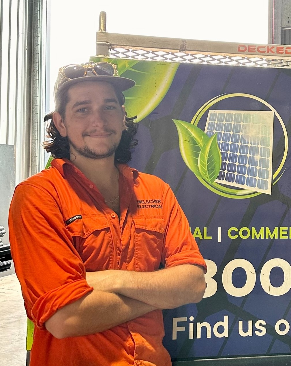 A young solar electrician named Harry in an orange work shirt and a cap stands smiling beside a team banner advertising solar panels.