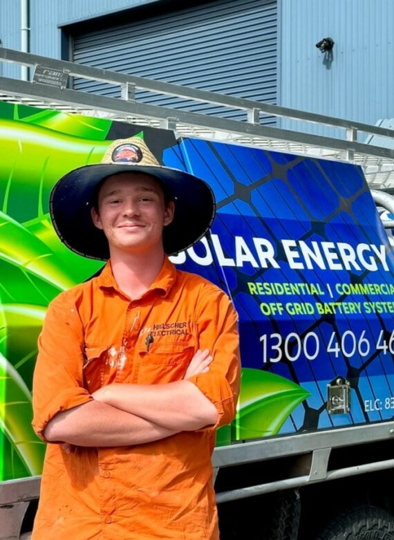 A young man in an orange shirt and wide-brimmed hat stands confidently in front of a colorful team solar energy service truck.