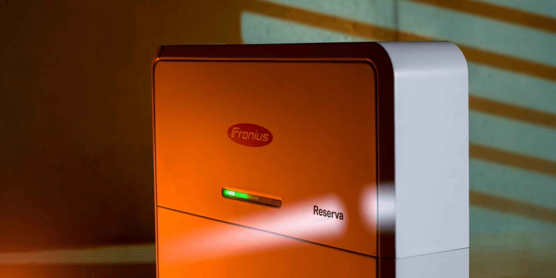 The orange and white "Fronius Reserva Cairns" device, labeled prominently, features a small display light, set against a backdrop of angled shadow lines. It's an integral part of advanced solar battery systems.