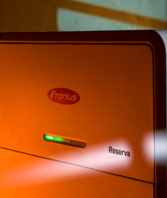 The orange and white "Fronius Reserva Cairns" device, labeled prominently, features a small display light, set against a backdrop of angled shadow lines. It's an integral part of advanced solar battery systems.