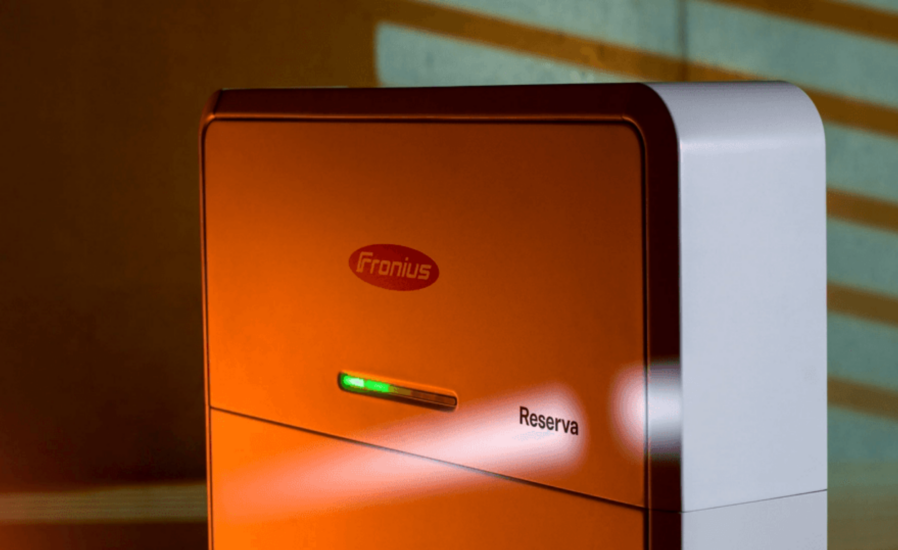 The orange and white "Fronius Reserva Cairns" device, labeled prominently, features a small display light, set against a backdrop of angled shadow lines. It's an integral part of advanced solar battery systems.