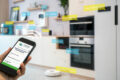 A person holds a smartphone displaying an appliance efficiency certification; in the background, various smart appliances in Cairns are labeled, including an oven, refrigerator, and more.