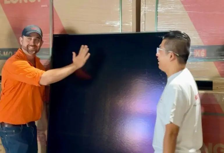 In Cairns, two men stand in front of stacked boxes, with one man in an orange shirt gesturing towards a large black Longi solar panel.