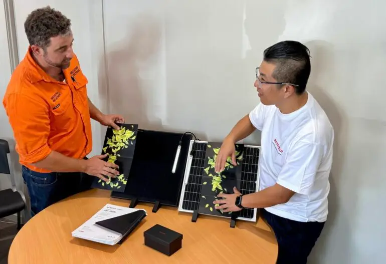 Two men inspect Longi Solar Cairns portable solar panels, which have leaves scattered on them, arranged on a round table in Cairns—surrounded by documents, a small box, and a pen.
