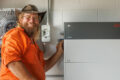 In Cairns, a man wearing an orange shirt and hat stands indoors, adjusting a battery storage unit mounted on the wall beside other electrical equipment.