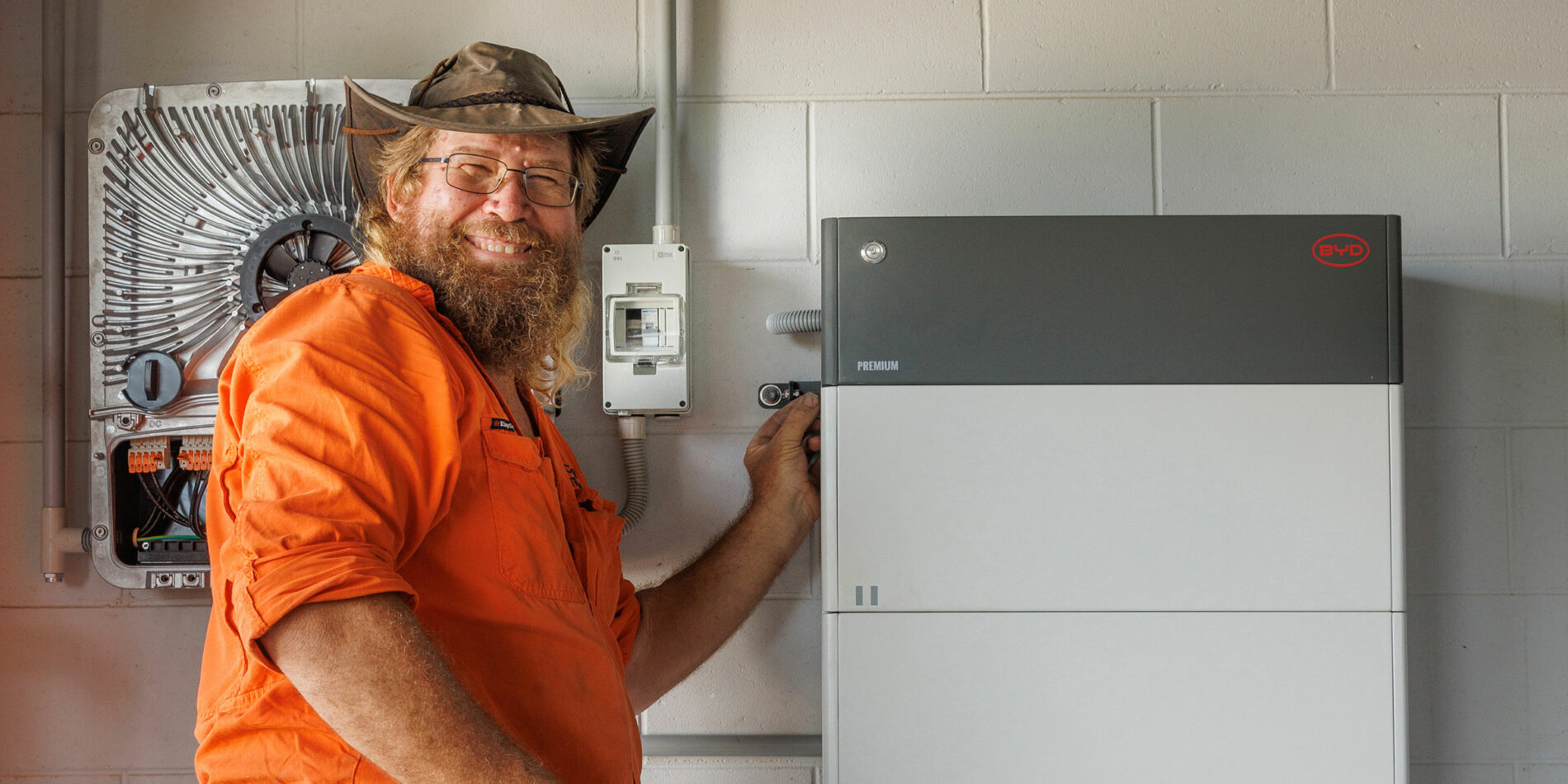 In Cairns, a man wearing an orange shirt and hat stands indoors, adjusting a battery storage unit mounted on the wall beside other electrical equipment.