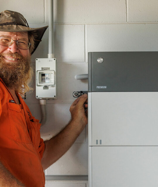 In Cairns, a man wearing an orange shirt and hat stands indoors, adjusting a battery storage unit mounted on the wall beside other electrical equipment.