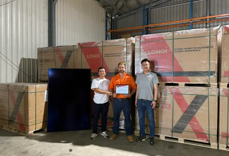 Three men stand in a warehouse in Cairns, surrounded by stacked boxes labeled "LONGi." The man in the center holds a certificate, showcasing the Longi Solar Cairns partnership, while the others stand beside him.