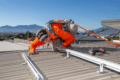 In Cairns, a worker clad in safety gear installs metal rails—essential components of residential solar systems—on a sloped roof beneath the clear blue skies typical of Far North Queensland. Tools and solar panels can be seen in the background, completing the scene.