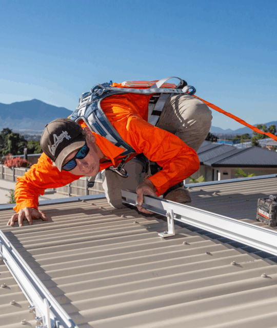 In Cairns, a worker clad in safety gear installs metal rails—essential components of residential solar systems—on a sloped roof beneath the clear blue skies typical of Far North Queensland. Tools and solar panels can be seen in the background, completing the scene.