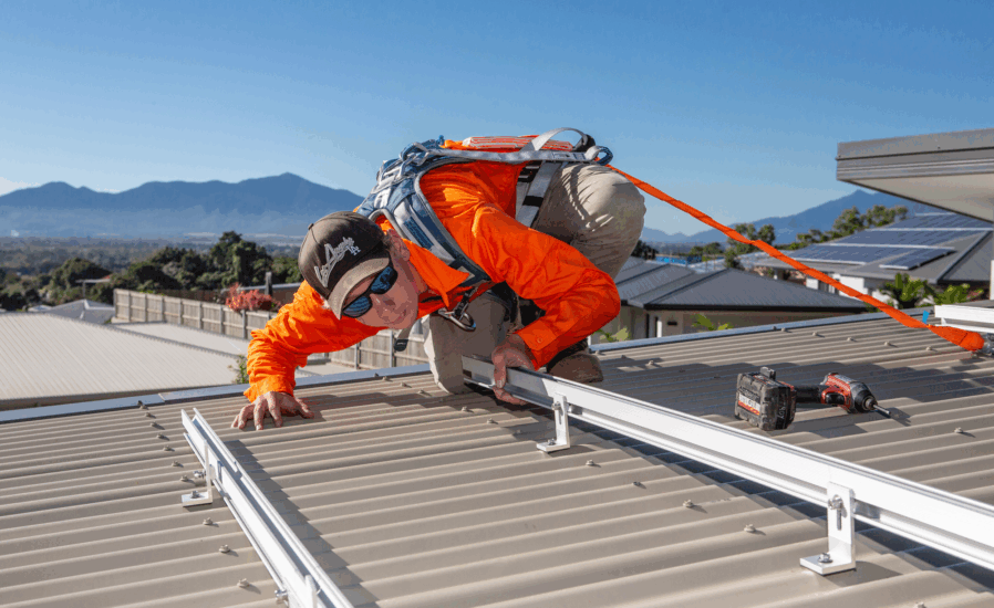 In Cairns, a worker clad in safety gear installs metal rails—essential components of residential solar systems—on a sloped roof beneath the clear blue skies typical of Far North Queensland. Tools and solar panels can be seen in the background, completing the scene.